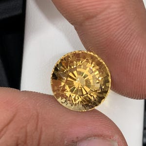 May include: A round, faceted gemstone with a golden-yellow hue. The gem is held between two fingers, showcasing its intricate cut and brilliance. The gemstone's facets reflect light, creating a sparkling effect. The gem is likely a citrine or a similar type of gemstone.