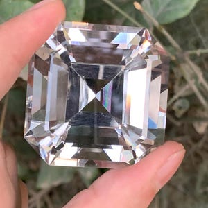 May include: A large, clear, square-cut gemstone with multiple facets, held between fingers. The gemstone reflects light, creating a rainbow effect. The background is blurred, with green foliage visible.