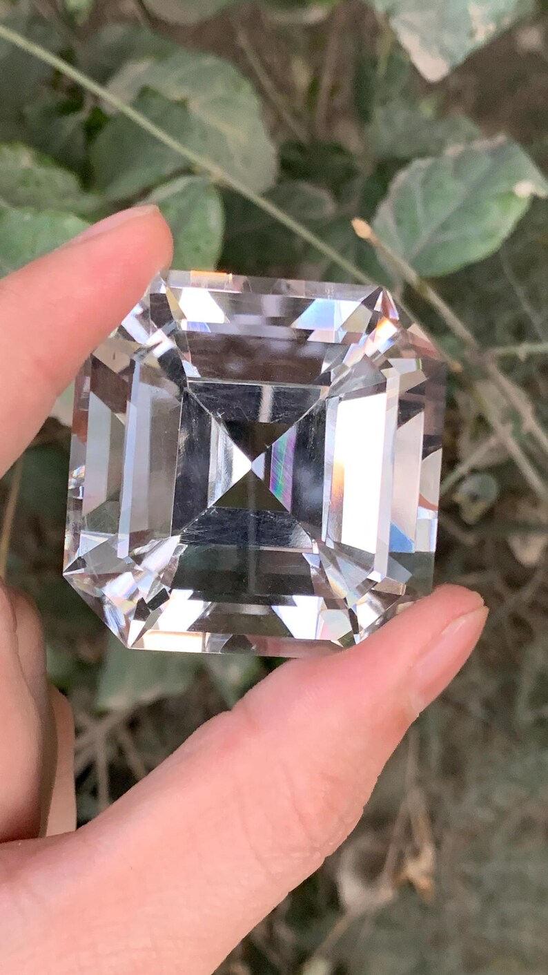May include: A large, clear, square-cut gemstone with multiple facets, held between fingers. The gemstone reflects light, creating a rainbow effect. The background is blurred, with green foliage visible.