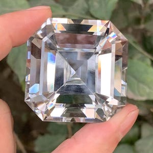 May include: A large, clear, square-cut gemstone with multiple facets, held between fingers. The gemstone reflects light, creating a rainbow effect. The background is blurred green foliage.
