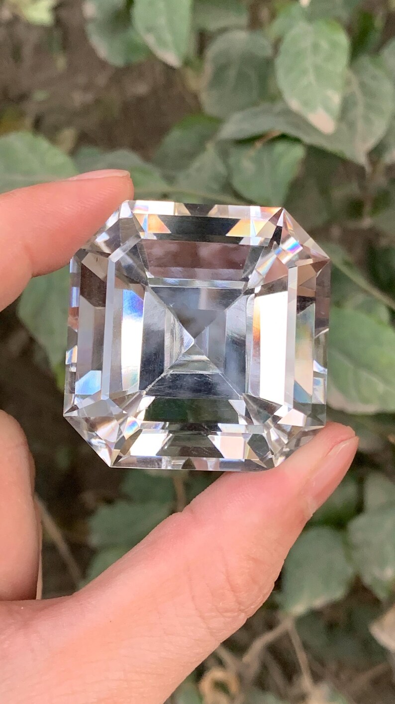 May include: A large, clear, square-cut gemstone with multiple facets, held between fingers. The gemstone reflects light, creating a rainbow effect. The background is blurred green foliage.