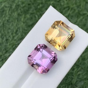May include: Two square-cut gemstones, one golden yellow and one pink, are presented on a white surface. The gems reflect light, and the background is a blurred green. The gems are cut with square corners.