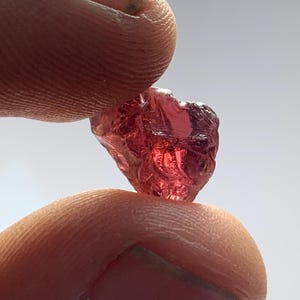 Op de afbeelding: Een ruwe, gefacetteerde, rode edelsteen die tussen twee vingers wordt gehouden. De edelsteen heeft een kristallijne structuur en reflecteert licht, waardoor zijn natuurlijke, ruwe schoonheid wordt getoond. De kleur van de steen varieert van diep rood tot een lichtere, doorschijnende tint.