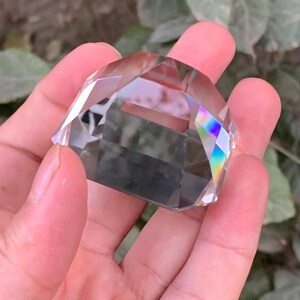 May include: A clear, faceted crystal gemstone held in a hand. The crystal has multiple flat surfaces and reflects a rainbow of colors. The background is blurred green foliage.