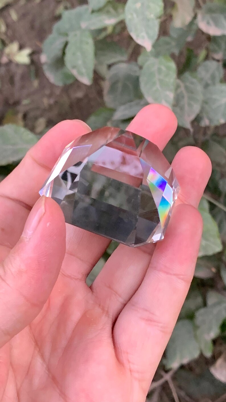 May include: A clear, faceted crystal gemstone held in a hand. The crystal has multiple flat surfaces and reflects a rainbow of colors. The background is blurred green foliage.
