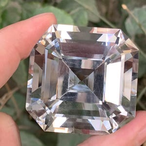 May include: A large, clear, square-cut gemstone with a multi-faceted design. The gemstone is held between fingers against a blurred green background. The cut of the stone creates a play of light and shadow, enhancing its brilliance.