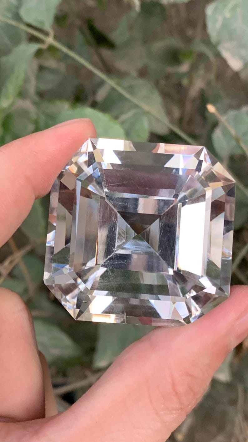 May include: A large, clear, square-cut gemstone with a multi-faceted design. The gemstone is held between fingers against a blurred green background. The cut of the stone creates a play of light and shadow, enhancing its brilliance.