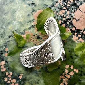 May include: A silver spoon ring with a floral design. The ring is bent into a unique shape and is sitting on a green and pink background.