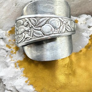 Puede incluir: Un anillo de plata hecho con una cuchara reciclada, con un diseño grabado de un pájaro y fruta. El anillo tiene un aspecto único y rústico.