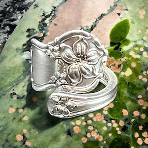 Puede incluir: Un anillo de cuchara de plata con un diseño floral. El anillo está hecho de una cuchara vintage y presenta una flor grande con flores más pequeñas que la rodean.