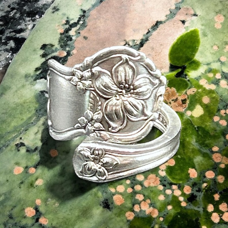May include: A silver spoon ring with a floral design. The ring is made from a vintage spoon and features a large flower with smaller flowers surrounding it.