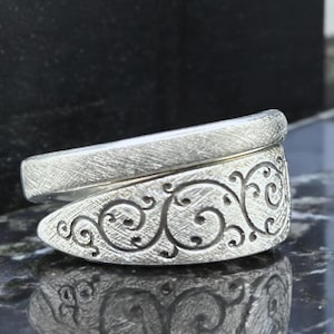 Puede incluir: Un anillo plateado con un patrón ornamental y arremolinado en la banda. El anillo tiene un acabado texturizado y un diseño único y superpuesto. La banda presenta un diseño de volutas grabado y detallado, añadiendo un toque de elegancia.