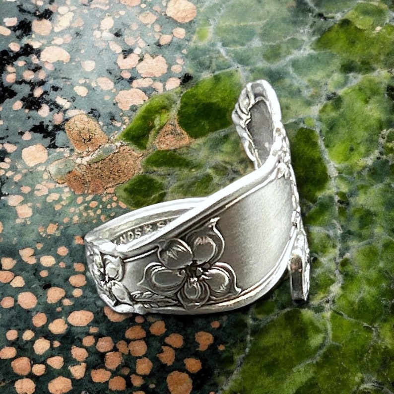 May include: A silver spoon ring with a floral design. The spoon is bent into a ring shape and has a flower design on the front. The ring is on a green and brown background.