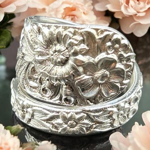 May include: A silver-colored ring with a floral design. The ring features detailed carvings of flowers and leaves. The ring is set against a backdrop of soft pink flowers, creating a delicate and elegant aesthetic.