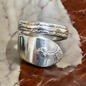May include: A silver spoon ring with an ornate design. The ring is made from a bent spoon and has a floral pattern.