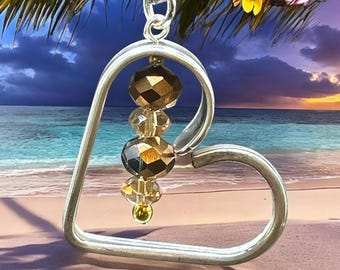 May include: A silver heart-shaped pendant necklace with a chain. The pendant has a clear crystal with gold accents hanging from it. The necklace is photographed against a beach scene with a palm tree and a sunset.