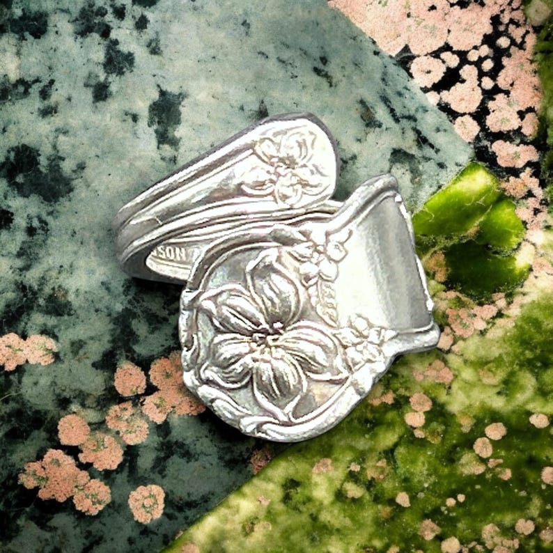 May include: A silver ring made from a repurposed spoon with a floral design. The ring is twisted and features a large flower with smaller flowers surrounding it.