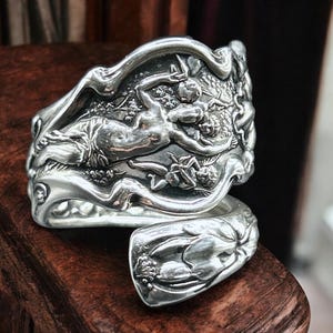 May include: A silver spoon ring with an ornate design featuring a reclining figure and cherubs. The ring is crafted from a spoon, with the spoon's bowl forming the ring's band. The detailed relief work showcases a classical theme.