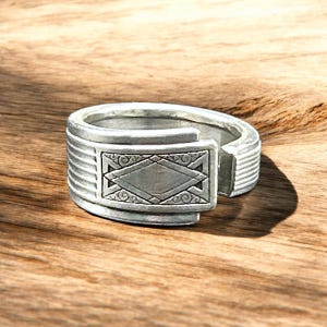 May include: A silver ring with a geometric design on the top. The ring is made of metal and has a textured surface.