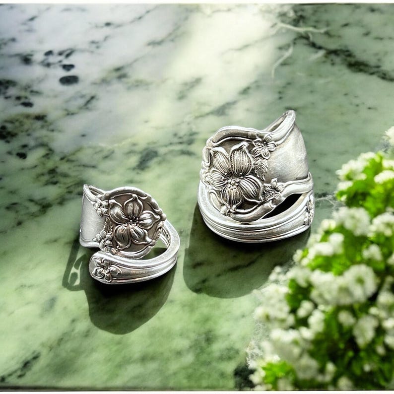 May include: Two silver rings made from repurposed silverware with a floral design. The rings are on a green marble surface.