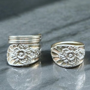 May include: Two silver rings with floral designs. The rings are made from repurposed silverware, featuring detailed flower patterns. One ring has a stacked band design, while the other has a simpler band. The rings are displayed on a gray surface.