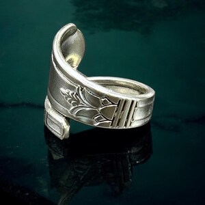 May include: A silver spoon ring with a decorative floral design. The ring is made from a repurposed spoon, with the spoon's bowl forming the top of the ring. The ring is set against a dark, reflective surface.