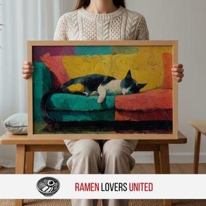 May include: A framed art print featuring a black and white cat sleeping on a green and red sofa. The artwork has a colorful, abstract background with yellow, orange, and pink hues. The frame is made of light-colored wood.