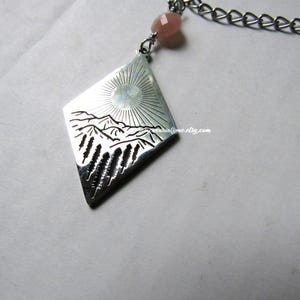 May include: Silver-toned pendant necklace with a diamond-shaped pendant featuring a mountain and sun design. The pendant is connected to a chain with a small pink bead. The necklace is a piece of jewelry.