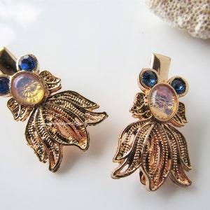 May include: Pair of gold-toned metal hair clips. Each clip features a filigree design resembling a goldfish, with an oval iridescent stone and two blue round stones. The clips have a barrette style clasp.