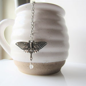 May include: Silver tea infuser featuring a detailed moth design and a clear glass bead at the end of the chain. The infuser is hanging from the rim of a white ceramic mug.