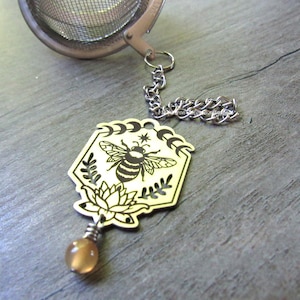 May include: A stainless steel tea infuser with a decorative charm. The charm features a laser-cut bee design with a lotus flower and moon phases. A small, round, amber-colored bead hangs from the charm.