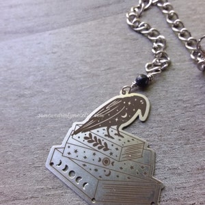 May include: A silver bookmark featuring a black crow perched on top of a stack of books. The books are decorated with stars, moons, and a sun. The bookmark has a silver chain attached to it.