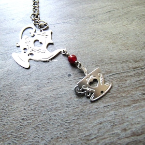 May include: A silver tea infuser featuring a teapot and teacup design, linked by a chain and a red bead. The teapot and teacup have heart-shaped cutouts and decorative scrollwork. The infuser is for steeping loose leaf tea.