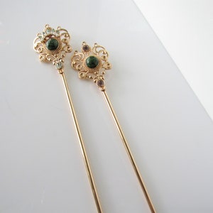 May include: Two gold hair sticks with ornate green and clear rhinestone embellishments. The sticks are long and slender, with a decorative design at the top.