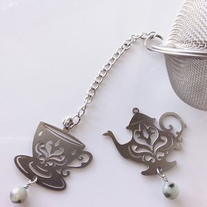 May include: Silver tea infuser with a teapot and teacup design. The infuser has a chain and a ball at the end. The ball is made of a light green stone.