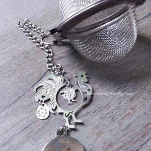 May include: Silver tea infuser with a rooster charm and a round charm with the text "Delicate Distraction" on it. The infuser has a chain and a mesh ball.