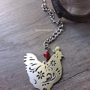 May include: Silver tea infuser with a chain and a red bead. The infuser is shaped like a rooster with a floral design.