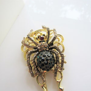 Puede incluir: Un broche de ara&ntilde;a de tono dorado con un cuerpo de piedras preciosas verdes y negras y detalles de diamantes de imitaci&oacute;n transparentes.