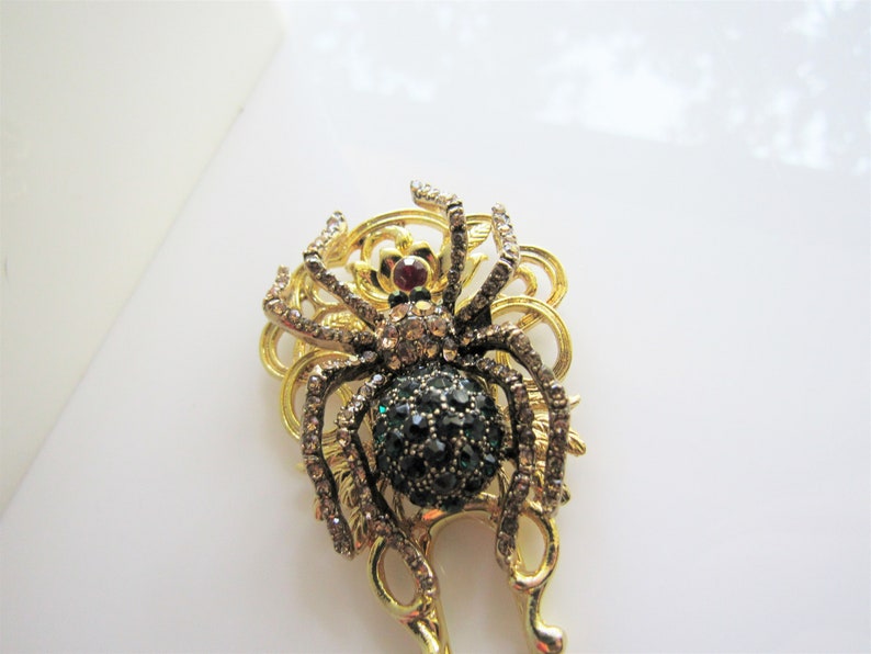 Puede incluir: Un broche de ara&ntilde;a de tono dorado con un cuerpo de piedras preciosas verdes y negras y detalles de diamantes de imitaci&oacute;n transparentes.