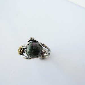 May include: A silver ring with a green and pink gemstone set in a twisted silver band. A small gold rose is on the side of the band.