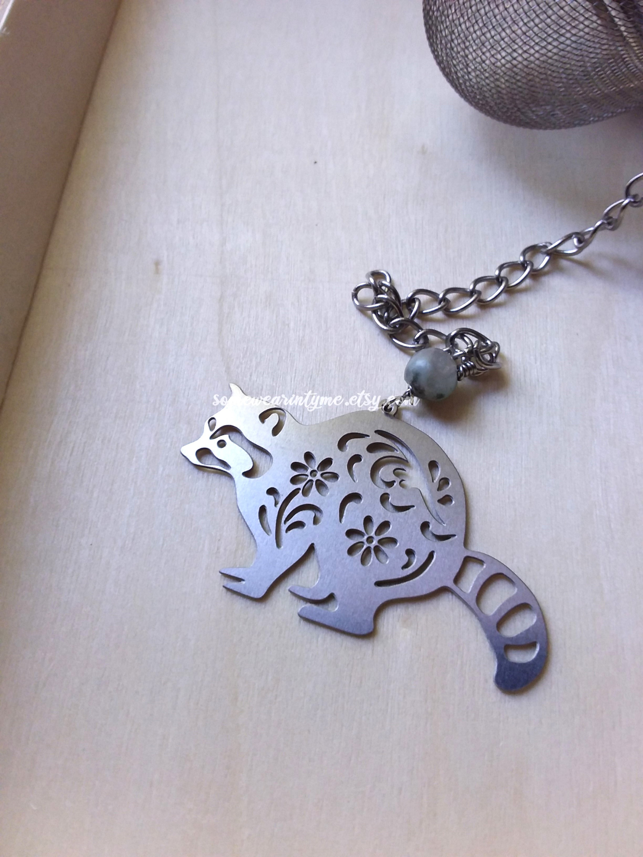 May include: Silver tea infuser shaped like a raccoon with floral cutouts. The infuser is attached to a chain with a green bead.
