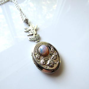 May include: Silver-toned oval locket necklace with a decorative floral design and a central opal-like stone. The locket is suspended from a delicate chain with a small charm. The necklace is a vintage-inspired piece.
