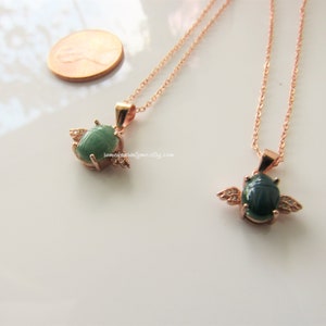 May include: Two gold necklaces with green scarab beetle pendants. The scarabs are detailed with white accents and have wings. The necklaces are on a white surface.