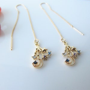 May include: A pair of gold dangle earrings with a crescent moon and stars design. The earrings are adorned with blue and white gemstones.
