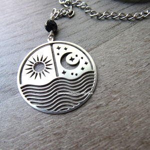 May include: Silver necklace with a round pendant featuring a sun, moon, and waves design. The pendant is attached to a silver chain with a black bead.