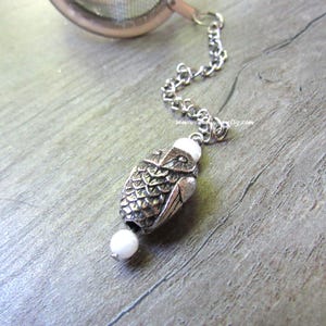 May include: A silver-toned tea infuser with a chain and an owl-shaped charm. The charm has a detailed, textured design and is accented with a small white bead at the bottom. The infuser is designed for steeping loose leaf tea.
