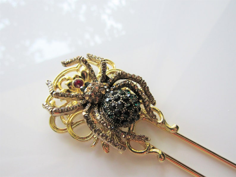 Puede incluir: Una varita para el pelo de tono dorado con un dise&ntilde;o de ara&ntilde;a decorativo. La ara&ntilde;a est&aacute; adornada con piedras de imitaci&oacute;n verdes y transparentes y una sola piedra de imitaci&oacute;n roja en el centro.