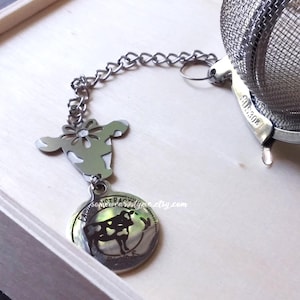 May include: Silver tea infuser with a chain and a cow charm. The charm has the text "Extraordinary" and a cow silhouette. The infuser has a mesh ball and a stainless steel handle.