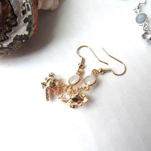 May include: Gold dangle earrings with a small fish charm and a white opal stone.
