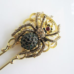 Puede incluir: Varilla para el pelo de tono dorado con un dise&ntilde;o de ara&ntilde;a adornada con gemas. La ara&ntilde;a tiene un cuerpo de gema verde y una gema roja en el centro de su cabeza. La ara&ntilde;a est&aacute; rodeada de un dise&ntilde;o de filigrana decorativo.